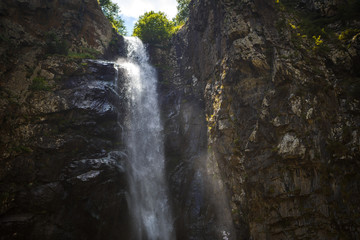 A high waterfall with white splashes of water descends from the mountain and sparkles in the sun. Attraction of Georgia Gveleti waterfall