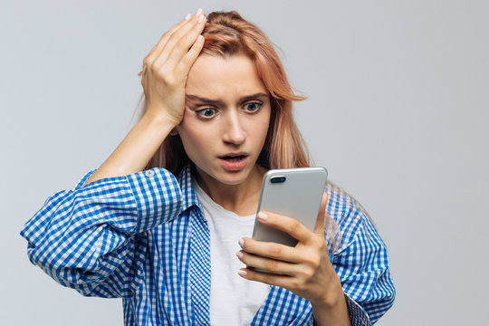 Studio portrait of young cute woman in checked shirt with facepalm gesture, touching head with palm, looking at phone. Epic fail, puzzled and worried face expression, reaction concept