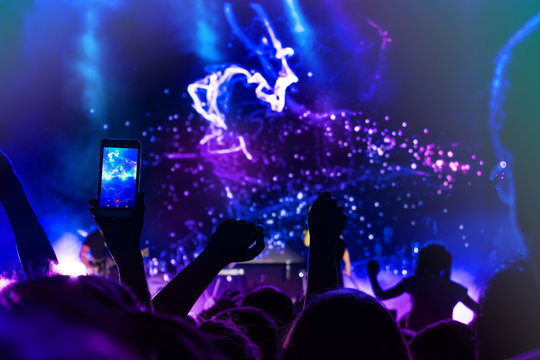 Crowd at concert. People silhouettes on backlit by bright blue and purple stage lights. Cheering crowd in colorful stage lights. Raised hands and smartphones against scene