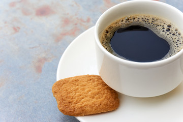 Simple white cup filled with black coffee on white saucer with brown biscuit on grey metal table with brown rust in soft light