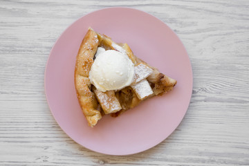Slice of homemade apple pie with ice cream on pink plate over white wooden background, top view. Flat lay, overhead, from above.