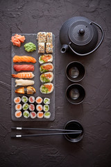 Assortment of different kinds of sushi rolls placed on black stone board. Traditional asian iron tea pot on side. Top angle view.