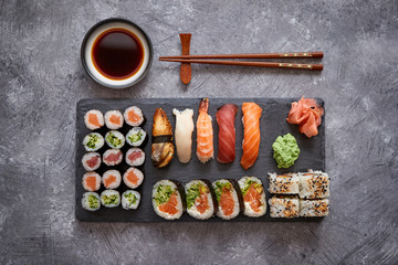 Composition of different kinds of sushi rolls placed on black stone board. Chopsticks and soy sauce bowl on side. Top angle view.