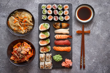 Japanese food composition. Various kinds of sushi placed on black stone board. Spicy kimchi salad, wontong soup, chopsticks and soy souce bowl.