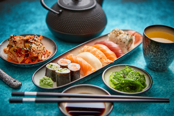 Various sushi rolls placed on ceramic plates. Traditional iron tea pot and green tea in cup. Kimchi and goma wakame salads. Soy souce and chopsticks on sides. Blue background.