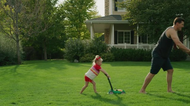 Daughter pushing toy lawnmower following father mowing lawn / Pleasant Grove, Utah, United States