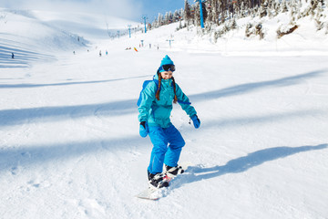 Fototapeta na wymiar A beautiful girl in winter clothes is riding a snowboard. She is wearing a blue helmet and a green jacket. having a great time in the mountains. Concept of travel, leisure, freedom, sport.