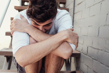 Depressed young adult man  hugging himself and sitting alone, mental illness health concept - obrazy, fototapety, plakaty