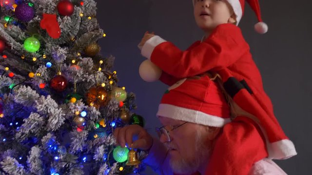 happy little child dressed in sainta claus suit is riding on shoulders of daddy deer image around Christmas tree. Family prepare and rehearse festive show. Christmas Eve. Happy New Year 2019