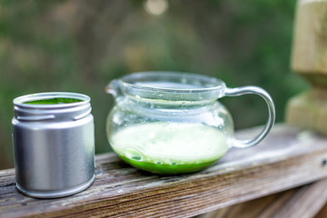 Glass teapot on wooden railing outside on backyard deck garden, filled with Japanese vibrant green tea color matcha, foam in morning