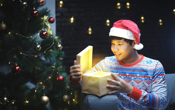 Asian Man With Christmas Costume Opening Shining Gift, Sitting Beside Christmas Tree