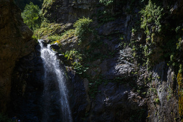 Fototapeta premium A high waterfall with white splashes of water descends from the mountain and sparkles in the sun. Attraction of Georgia Gveleti waterfall