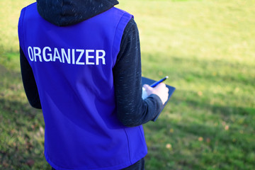 Organizer written on blue vest back of young man participates as volunteer and writing results outdoor sport event. Blurred green grass background with copy space for text.