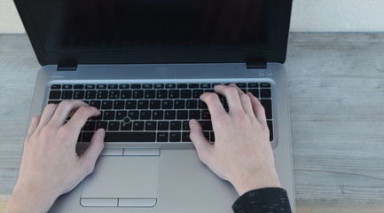 Hands typing keyboard of gray laptop with black screen on natural wood background.