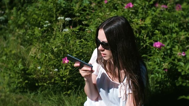 Beautiful brunette woman records a voice message on her mobile phone while sitting in the park on a Sunny day.