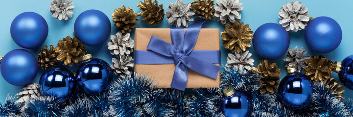 Gift box with blue ribbon, Christmas-tree decorations, balls and white and gold pine cones on a blue background. Concept of Merry Christmas and Happy New Year. Minimalism. Flat lay, top view. Banner