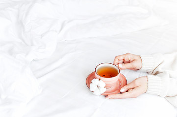 Fototapeta na wymiar Women hands in white knitted sweater hold cup of hot tea on saucer with delicate cotton flower in the bed. Cozy morning breakfast at home. Lifestyle gentle background Copy Space autumn winter concept