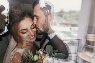 Gorgeous bride and stylish groom gently hugging at window. Happy wedding couple embracing and smiling. Romantic moments of newlyweds. Creative wedding photo. Copy space