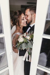 Gorgeous bride and stylish groom gently kissing at window. Happy wedding couple embracing. Romantic...