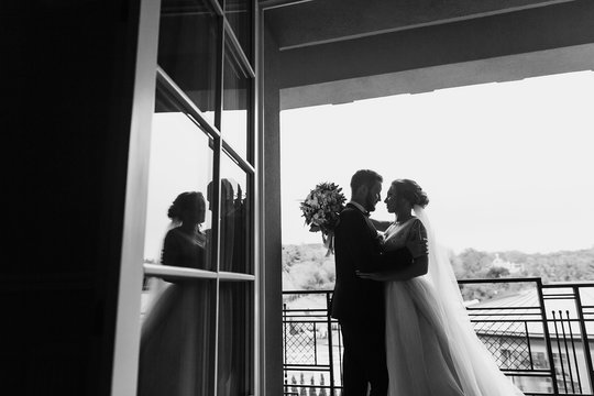 Gorgeous bride and stylish groom silhouettes at window light. Sensual wedding couple embracing. Romantic moments of newlyweds. Creative wedding photo. Copy space