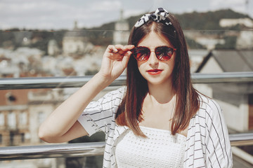 Beautiful stylish young woman in  sunglasses, retro dress and headband relaxing on roof in european city street. Hipster girl smiling and enjoying summer time in Lviv. Space for text