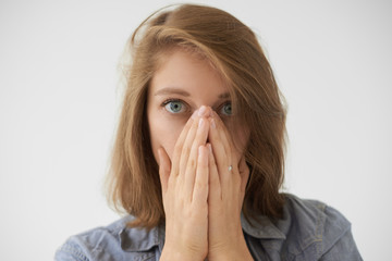 Close up picture of frightened young woman with loose hairdo covering face with both hands and staring at camera with scared look, being stressed or terrified with something. Human emotions