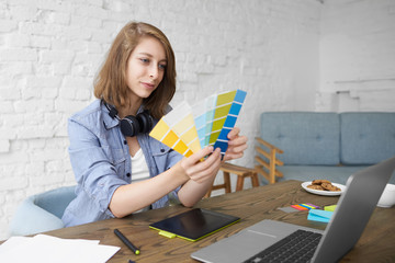 Portrait of positive young stylish woman designer sitting at desk in front on open laptop, showing...