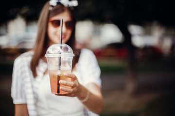 Ban single use plastic. Happy hipster girl with sunglasses, in retro dress, holding plastic cup and straw. Beautiful stylish young woman holding cold drink and relaxing in city street.