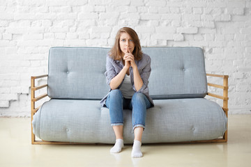 Concerned focused young woman in jeans and white socks sitting on grey couch against white brick...