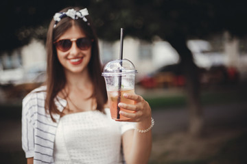 Beautiful stylish young woman holding cold drink and relaxing in city street. Happy hipster girl with sunglasses, in retro dress, holding single use plastic cup and straw. Ban plastic