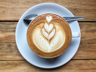  Hot cappuccino coffee in white cup and saucer with spoon on wooden table background. Art of milk foam drawing like white heart on brown melt coffee top view