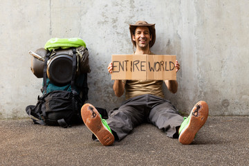 A single white adult man with a packed backpack sitting on the roadside in front of a gray wall wearing broken green shoes and holding a cardboard sign reading entire world