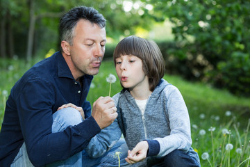 Handsome man with his son blowing dandelions over blurred green grass, summer nature outdoor. Father with liitle son playing in summer park. Parenting. Family.