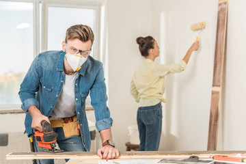 young man in protective workwear using electric jigsaw and looking at camera while wife painting wall behind