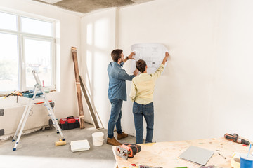 back view of young couple holding blueprint at wall during repairment