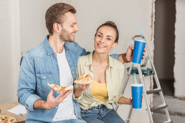 smiling young couple holding paper cups with drinking straws and pizza slices during repairment