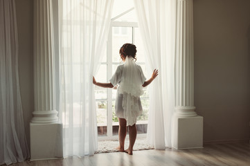 Gorgeous beautiful bride in silk robe looks out the window and waiting for her groom. Wedding morning