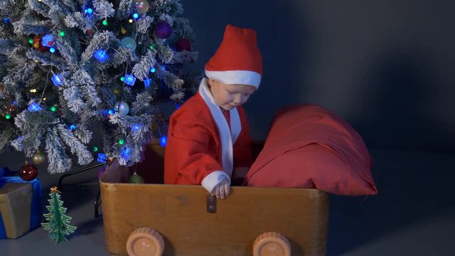 Cute boy dressed in a festive costume of Santa Claus is sleeping in a wooden box under the Christmas tree and waiting for gifts. Christmas Eve