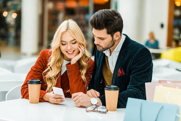 smiling couple using smartphone in cafe with coffee to go and shopping bags