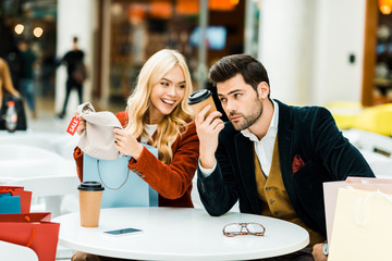 happy fashionable girl showing new clothes to confused boyfriend in cafe in shopping center