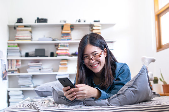 Pretty Asian Woman With Glasses Lying On The Bed Reading Good News On Line In A Smart Phone, Woman Lifestyle