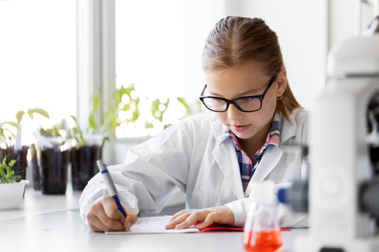 education, science and children concept - girl studying chemistry at school laboratory and writing to workbook