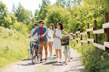 people, leisure and lifestyle concept - happy young friends with fixed gear bicycles walking along country road in summer