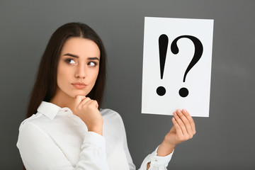 Thoughtful young woman holding sheet of paper with exclamation and interrogation marks on grey background