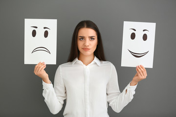 Emotional woman holding sheets of paper with drawn emoticons on grey background