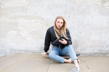 Teenage girl sitting on her skate board texting outside