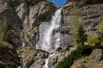 spring mountain waterfall, with green vegetation and snow on a blue sky
