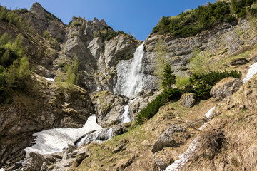 spring mountain waterfall, with green vegetation and snow on a blue sky
