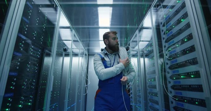 Medium shot of a male technician working in a data center carrying cable down the corridor amongst rows of server racks