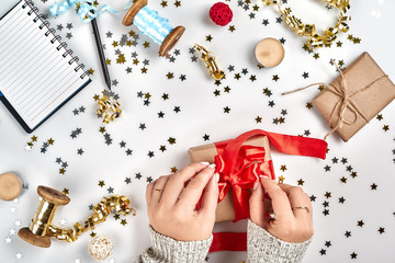 Female hands holding present with red bow on white background with delicate wavy ribbons and metallic star shaped confetti. Flat lay style
